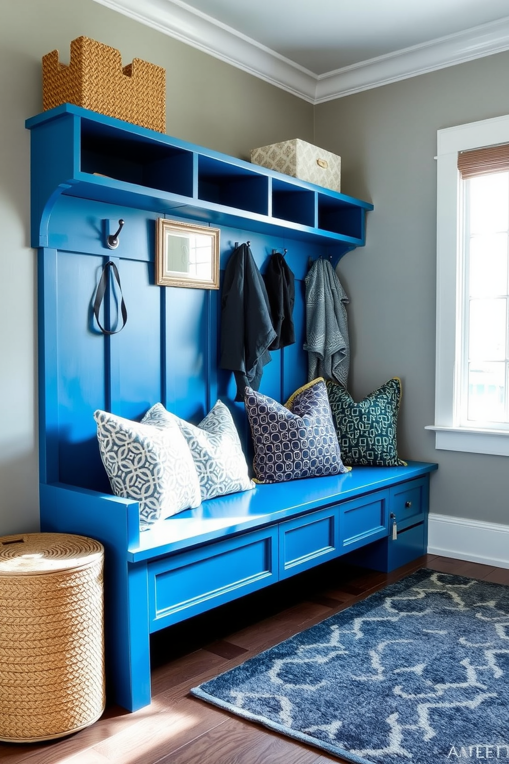 A sapphire blue bench is positioned against the wall, adorned with an array of patterned pillows in complementary colors. The mudroom features a combination of open shelving and hooks for coats, creating an inviting and functional space. The walls are painted in a soft gray hue, enhancing the brightness of the room. A stylish area rug anchors the space, while natural light floods in through a nearby window, illuminating the carefully curated decor.