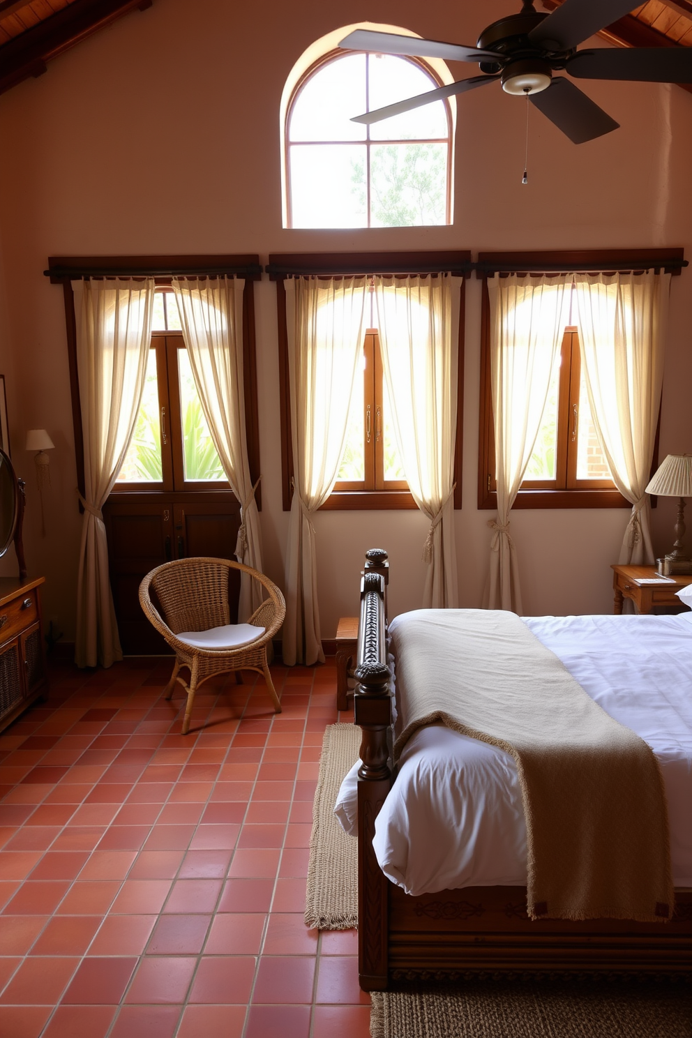 A warm Mediterranean master bedroom featuring terracotta tiles that add a rustic charm. The room is adorned with a large wooden bed framed by intricate carvings and topped with plush white linens. Natural light floods in through arched windows draped with sheer curtains, creating a soft and inviting atmosphere. A cozy seating area with woven chairs and a small wooden table sits in the corner, perfect for relaxation.