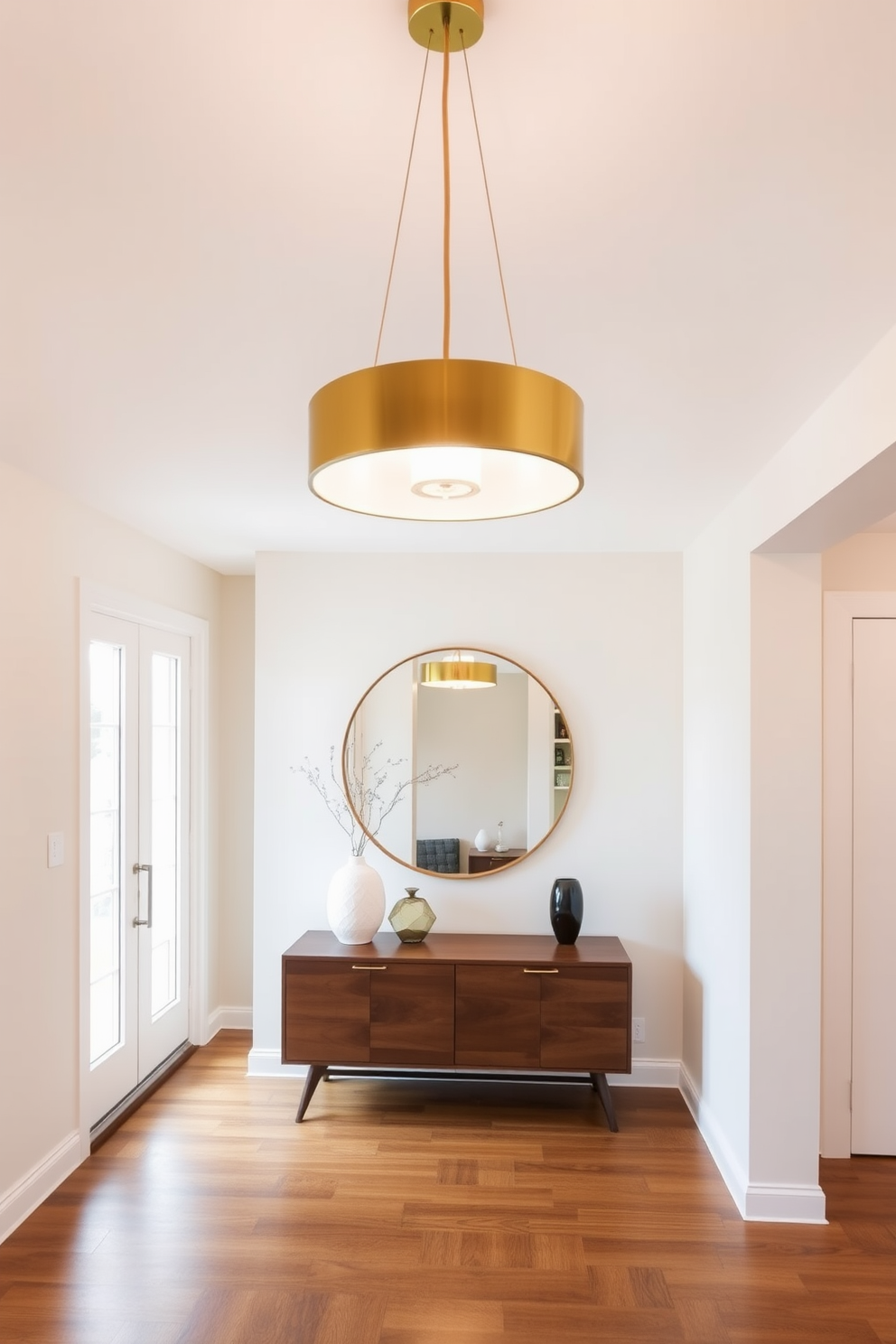 A stunning mid century modern pendant light fixture hangs gracefully from the ceiling, featuring a sleek design with a warm brass finish. Below it, a beautifully designed foyer welcomes guests, showcasing a minimalist console table adorned with a stylish vase and a geometric sculpture. The walls are painted in a soft, neutral tone that complements the rich wooden flooring, creating an inviting atmosphere. A large round mirror reflects the light fixture, enhancing the space's brightness and adding a touch of elegance.