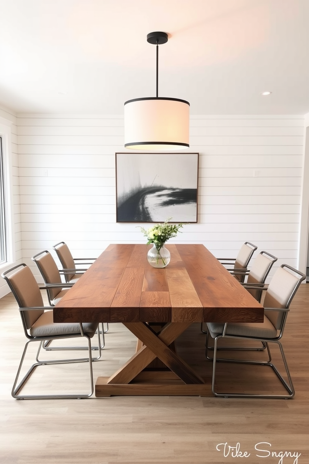 A rustic farmhouse table made of reclaimed wood sits at the center of the dining room. Surrounding the table are sleek modern chairs with metal frames and cushioned seats in a neutral fabric. The walls are adorned with shiplap in a soft white finish, creating a warm and inviting atmosphere. A large pendant light hangs above the table, casting a warm glow over the space and highlighting the natural textures.