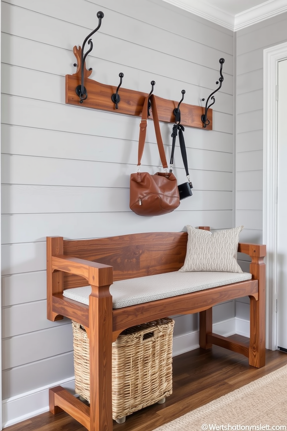 Artisan crafted bench featuring intricate woodwork and unique detailing. The bench is positioned against a backdrop of shiplap walls painted in a soft gray, with a woven basket underneath for storage. The seat is upholstered in a durable fabric with a subtle pattern, providing both comfort and style. Above the bench, a series of hooks are mounted for hanging coats and bags, enhancing the functionality of the space.