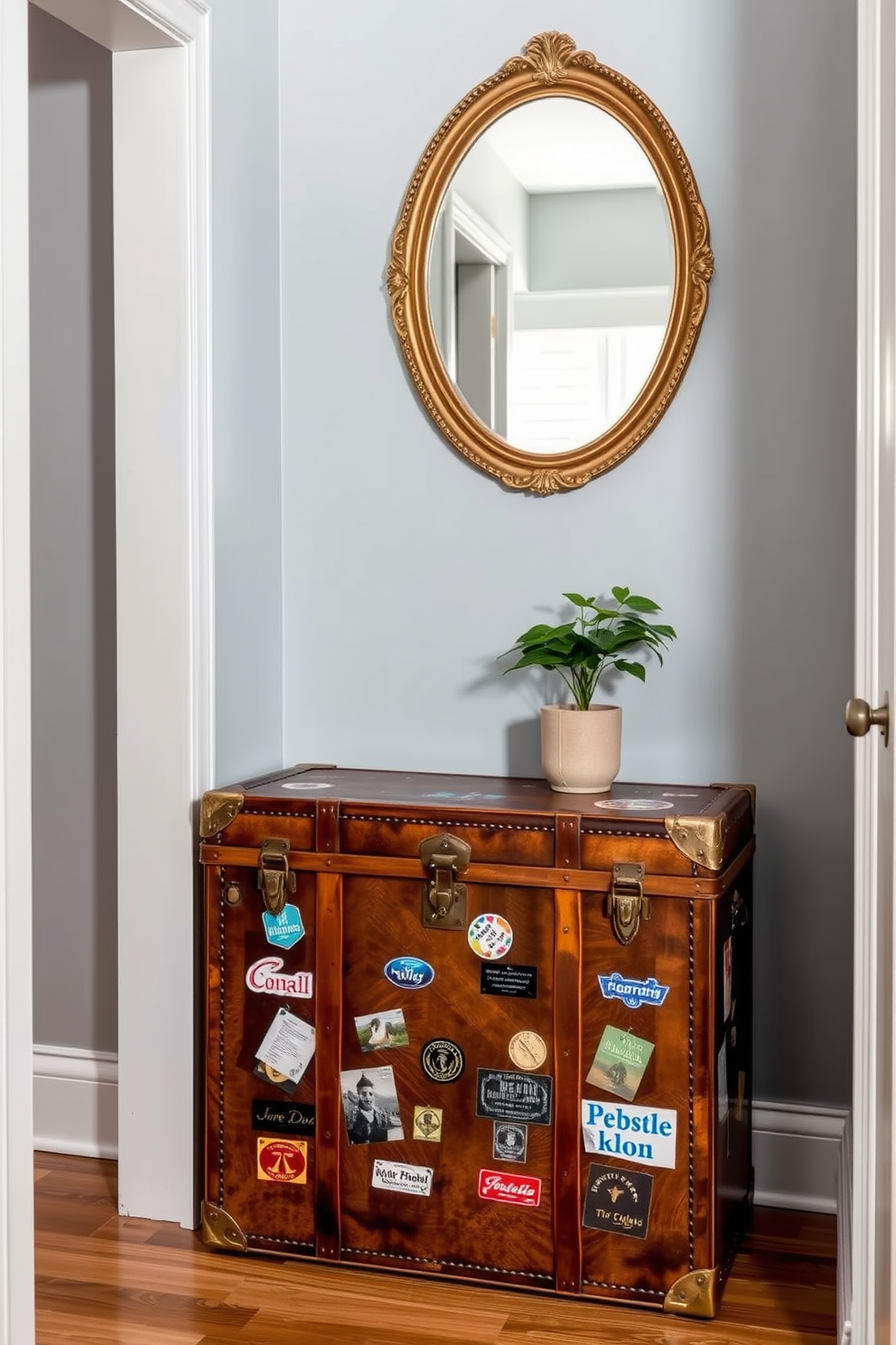 A vintage trunk serves as a unique storage solution in a narrow entryway. The trunk is adorned with travel stickers and sits against a wall painted in soft gray, complemented by a narrow console table above it for added functionality. The entryway features a round mirror with an antique gold frame that reflects natural light. A small potted plant rests on the console table, adding a touch of greenery to the space.