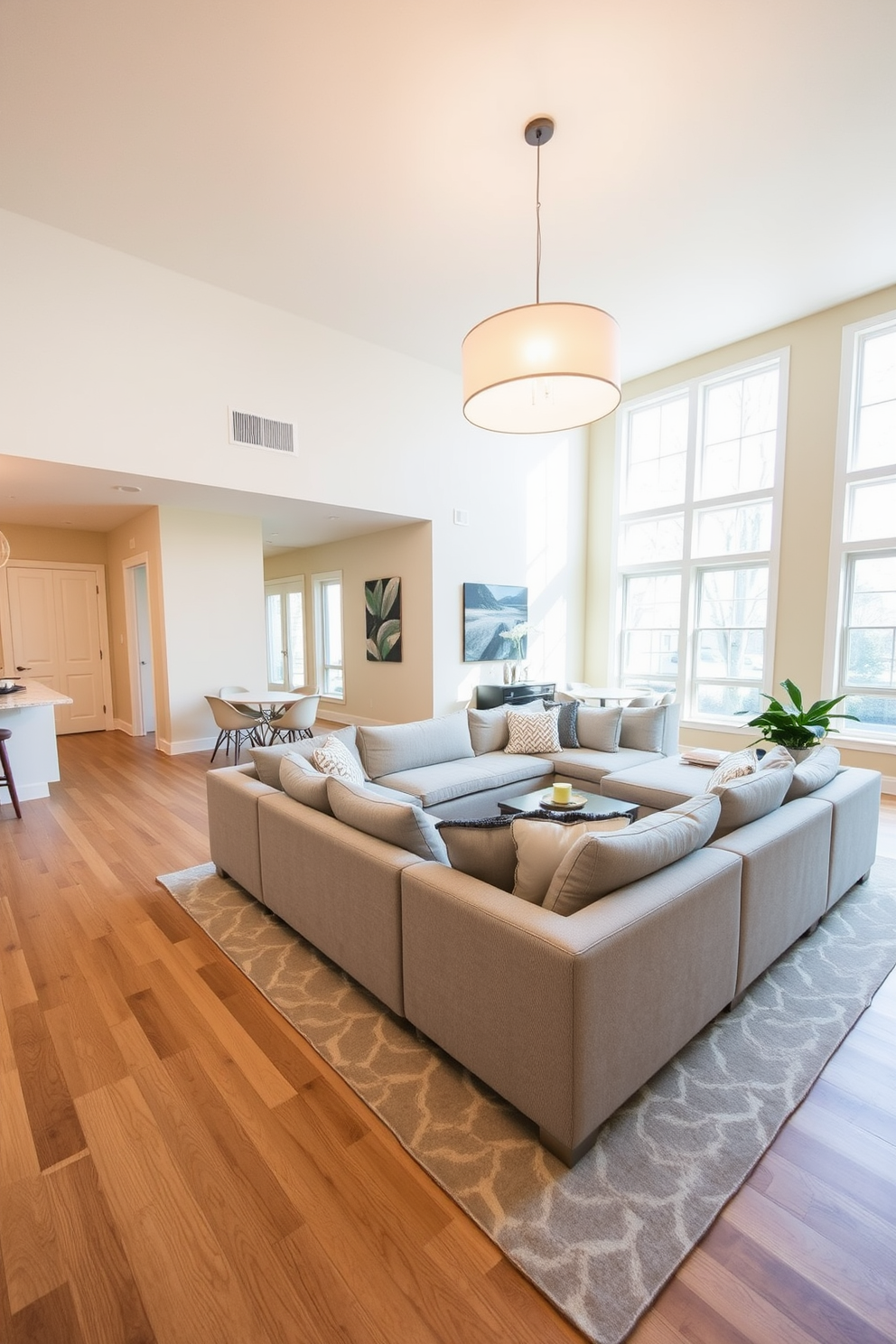 A spacious open floor plan living room features a comfortable sectional sofa in a soft gray fabric, complemented by a large area rug with subtle geometric patterns. The walls are painted in a warm cream tone, creating a welcoming atmosphere. A statement lighting fixture hangs overhead, casting a warm glow across the room. Large windows allow natural light to flood in, enhancing the airy feel of the space.