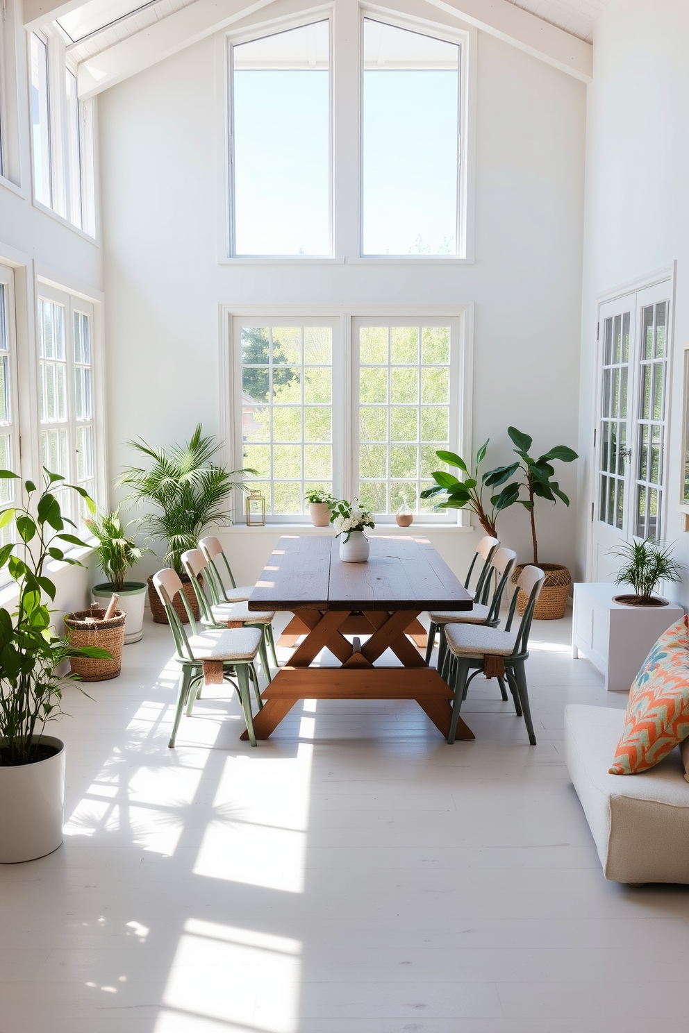 A bright and airy summer loft space features large windows that let in natural light. The room is adorned with light-colored furnishings and soft textiles to create a relaxed atmosphere. In the center, a rustic wooden picnic table serves as a dining option, surrounded by mismatched chairs for a casual vibe. Potted plants and colorful cushions add a touch of greenery and comfort to the space.