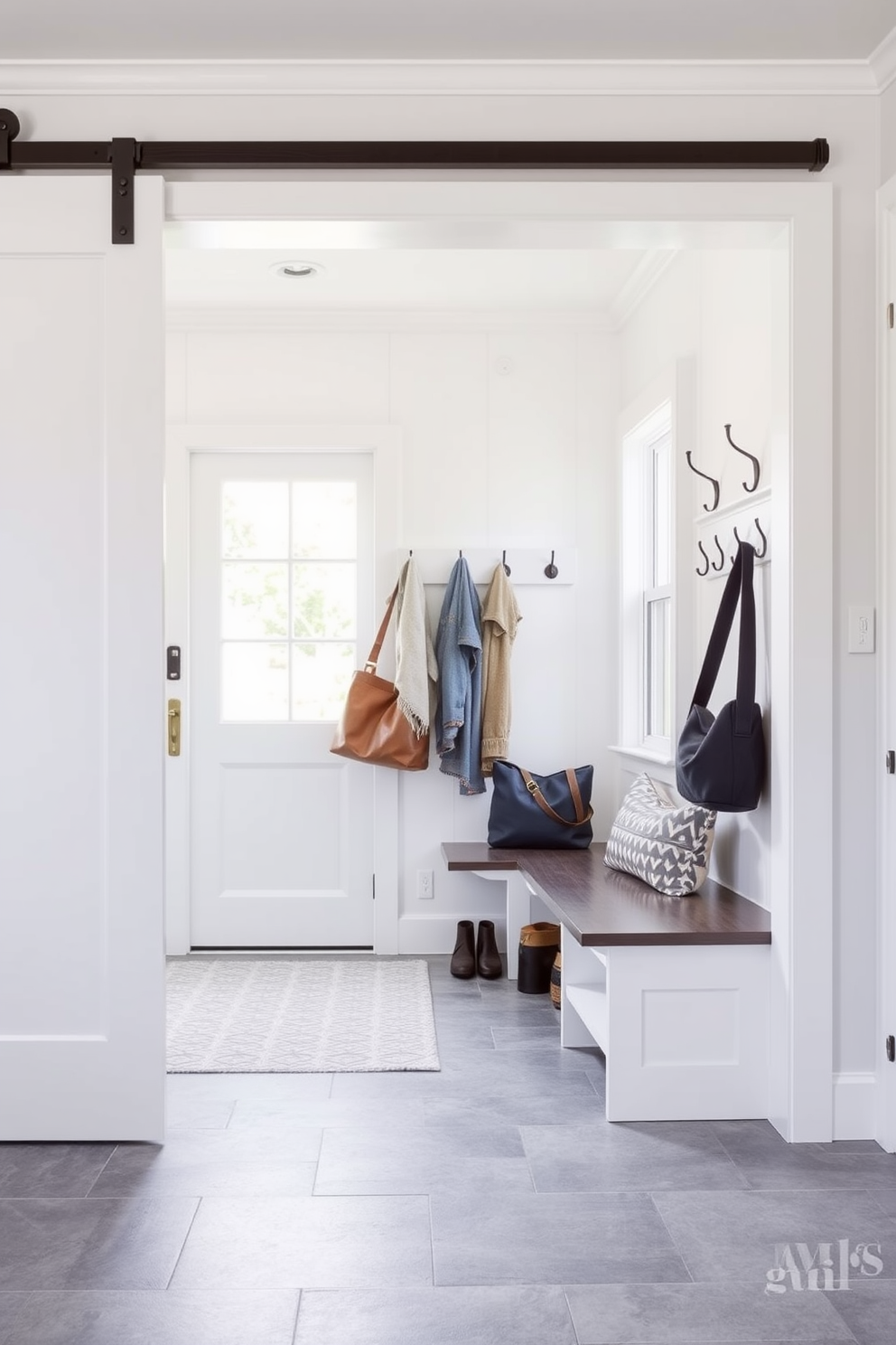 A chic mudroom featuring a sliding barn door opens into a bright and airy space. The walls are painted in a crisp white, complemented by a durable gray tile floor that adds a modern touch. Stylish hooks line the wall for coats and bags, while a built-in bench provides seating and storage underneath. A large window allows natural light to flood the room, enhancing the inviting atmosphere.